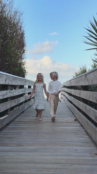 Girl wearing Indie Blue Sylvia Dress Blue Mist Floral and Boy wearing Ty Shirt White Linen with Finn Pants walking on boardwalk near beach