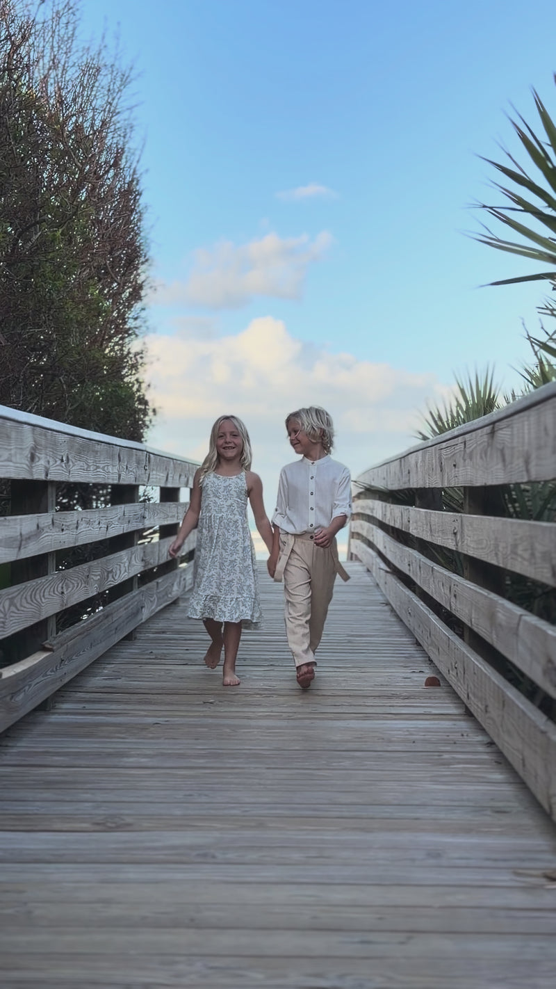 Girl wearing Indie Blue Sylvia Dress Blue Mist Floral and Boy wearing Ty Shirt White Linen with Finn Pants walking on boardwalk near beach